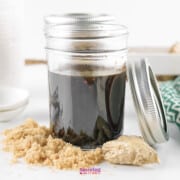 A jar of Teriyaki Sauce with a metal lid off to the side, accompanied by brown sugar and a cloth napkin on a light surface.