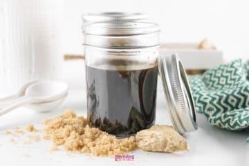 A jar of Teriyaki Sauce with a metal lid off to the side, accompanied by brown sugar and a cloth napkin on a light surface.