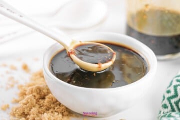 A spoon lifting Teriyaki Sauce from a small white bowl, with brown sugar and a jar visible in the background.