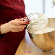 A person in a red chef's jacket holds a whisk dripping with white cream above a stainless steel mixing bowl.