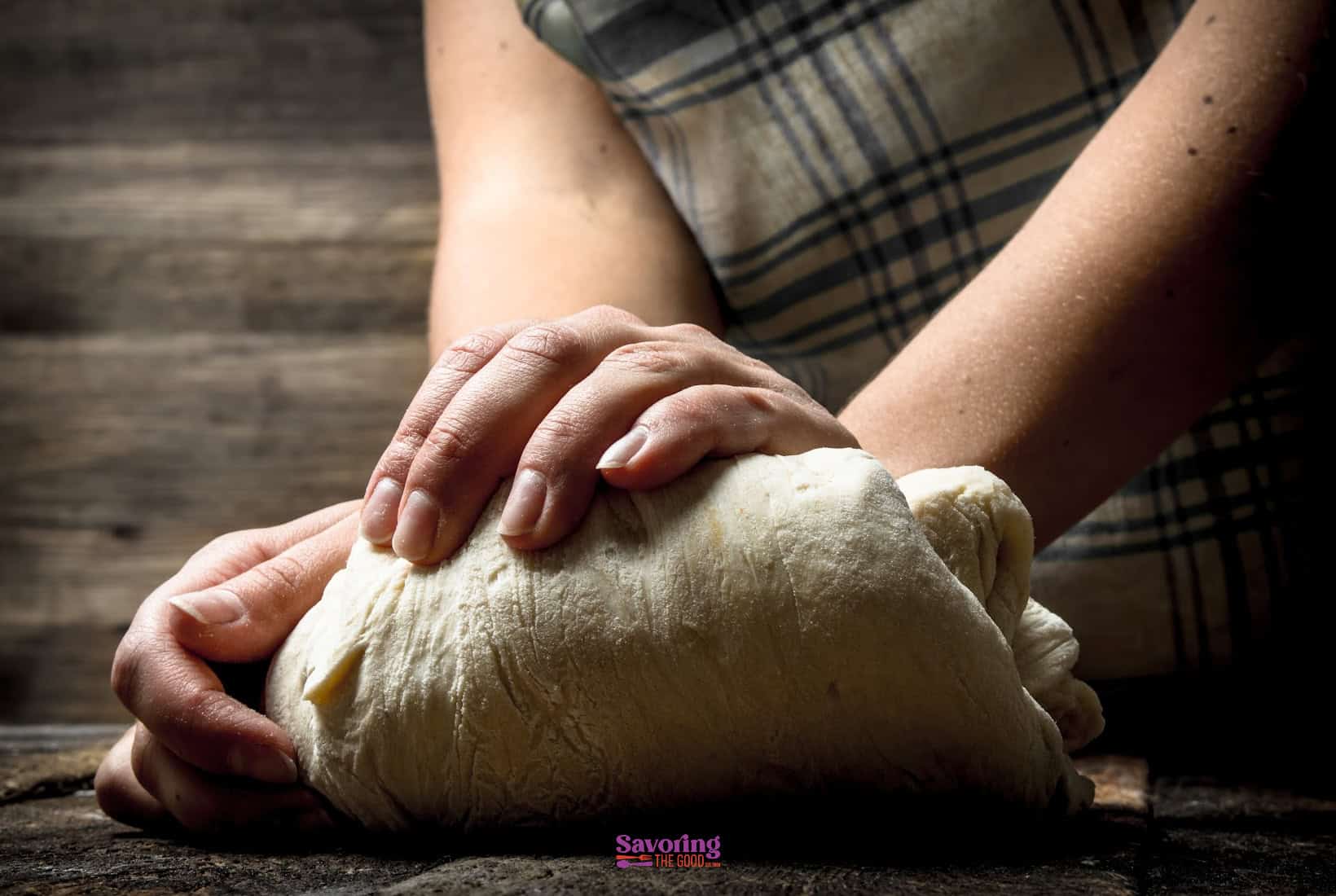 A person kneading a ball of dough on a wooden surface, wearing an apron, focusing on their hands pressing down on the dough.