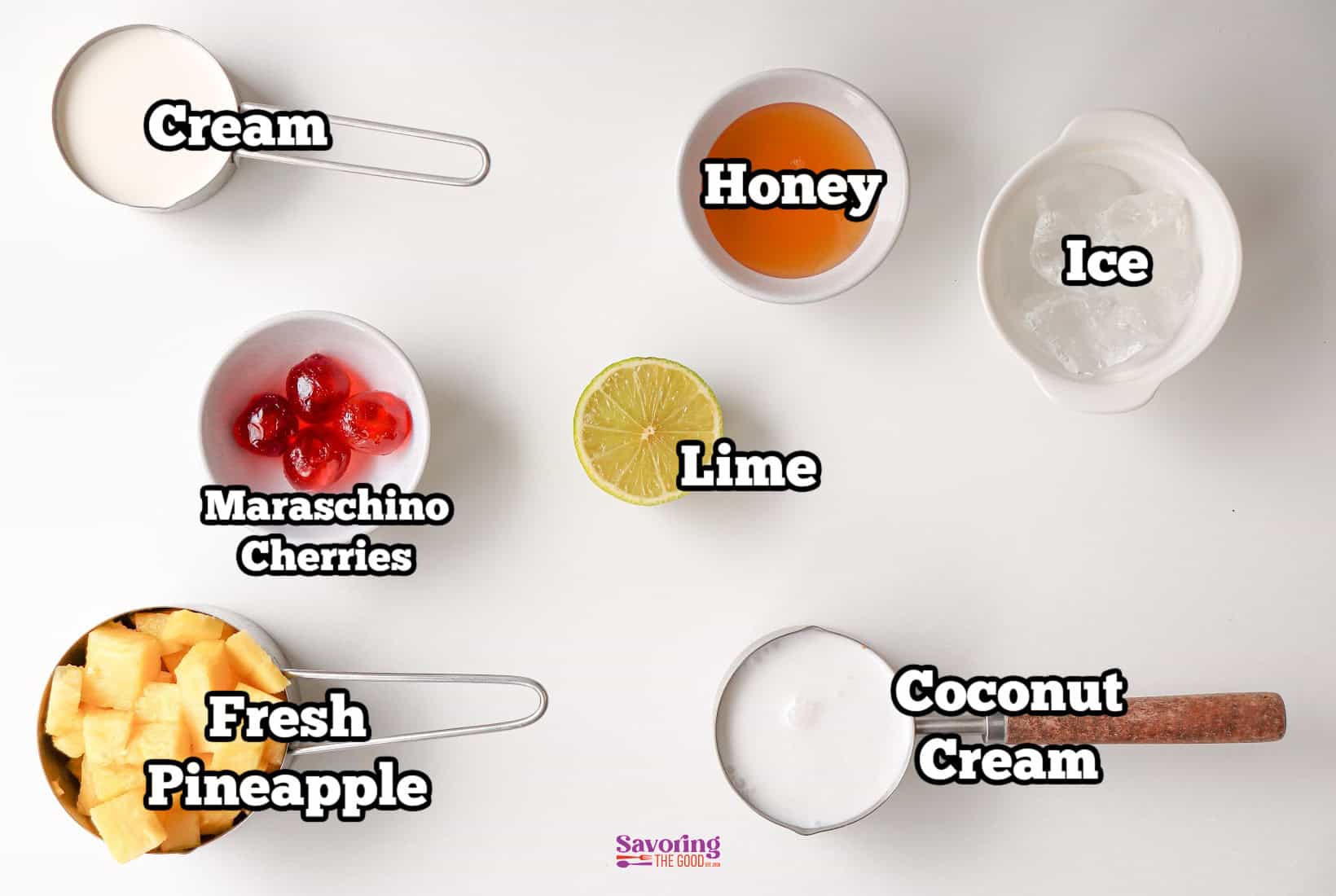 Top-down view of various ingredients for a pina colada mocktail labeled: cream, honey, ice, maraschino cherries, lime, fresh pineapple, and coconut cream on a white