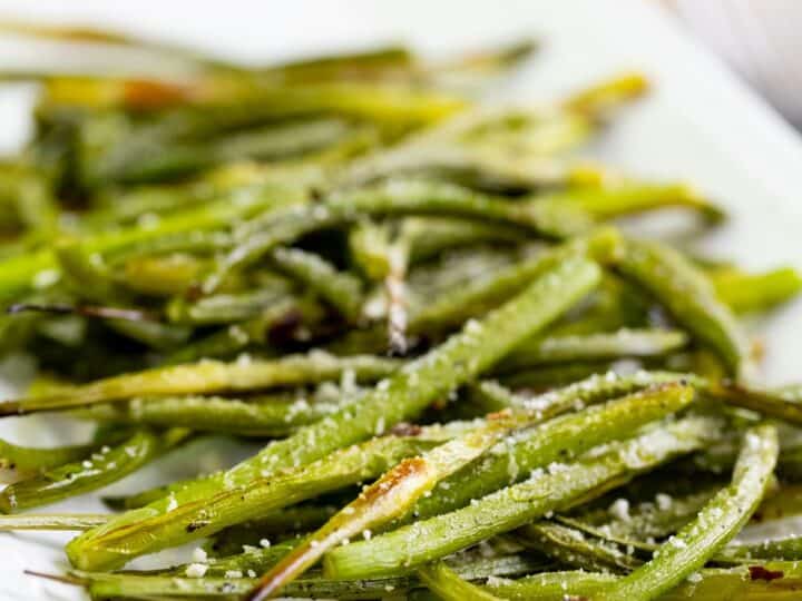 Roasted green beans and garlic scapes sprinkled with grated Parmesan cheese, served on a white rectangular plate.