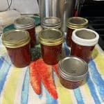 Seven jars filled with Strawberry Rhubarb Jam are placed on a striped towel, with lids of varying colors, in a cozy kitchen setting.