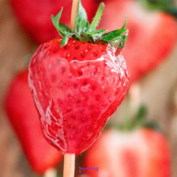 Close-up of a fresh Strawberry tanghulu on a stick, with a glossy appearance and other blurred strawberries in the background.