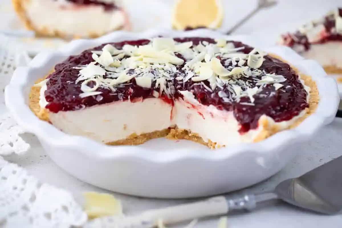 A sweet treat with a graham cracker crust, creamy white filling, topped with a layer of red fruit sauce and white chocolate shavings—one slice missing. Perfect for fans of no bake desserts—no heat required!.