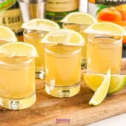 Five shot glasses filled with a light brown liquid, each garnished with a lime wedge, are arranged on a wooden board. Several lime wedges are placed alongside.