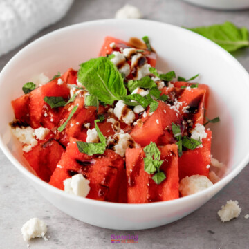A white bowl filled with watermelon cubes, topped with crumbled feta cheese, fresh mint leaves, and a drizzle of balsamic glaze.