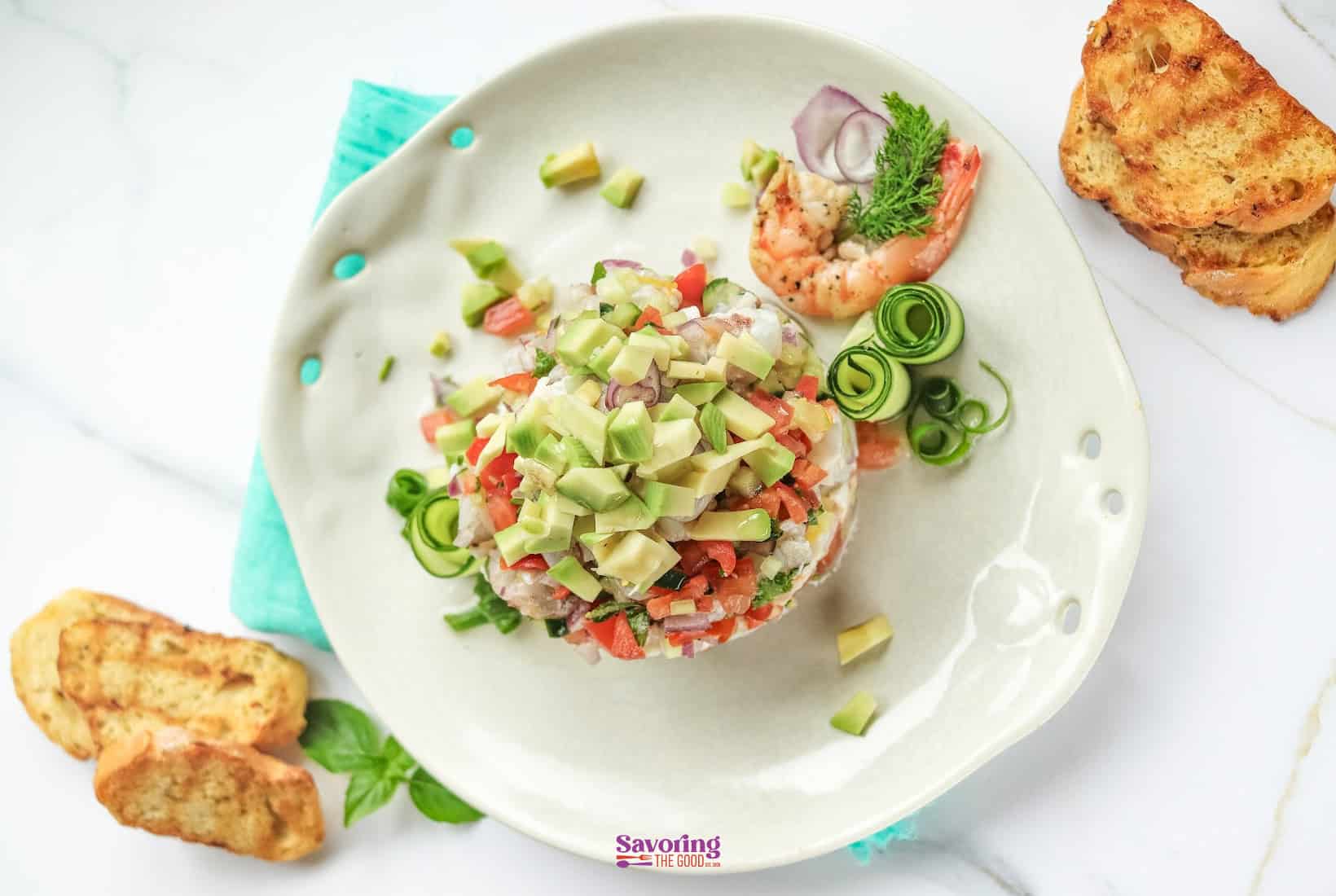 A round serving of shrimp ceviche topped with diced avocado and tomatoes, garnished with cucumber spirals and grilled bread on the side, presented on a white plate.