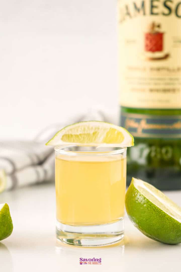 A shot glass filled with a yellow liquid, garnished with a lime wedge, sits in front of a partially visible bottle of Jameson whiskey. A slice of lime is placed on the side.