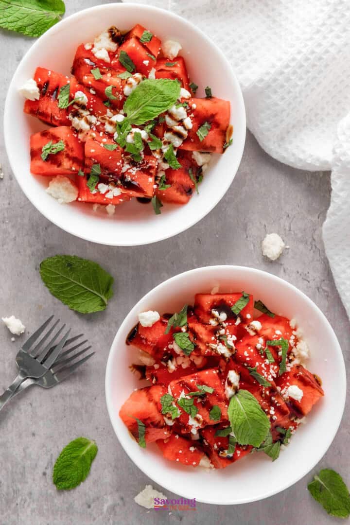 Two bowls of watermelon salad with feta cheese, fresh mint, and a drizzle of balsamic glaze on a gray surface with mint leaves scattered and a fork nearby.