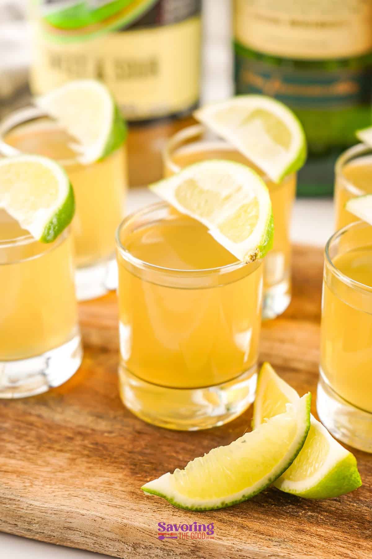 Six shot glasses filled with a light yellow liquid are garnished with lime wedges, placed on a wooden tray with additional lime wedges. Bottles in the background are slightly blurred.