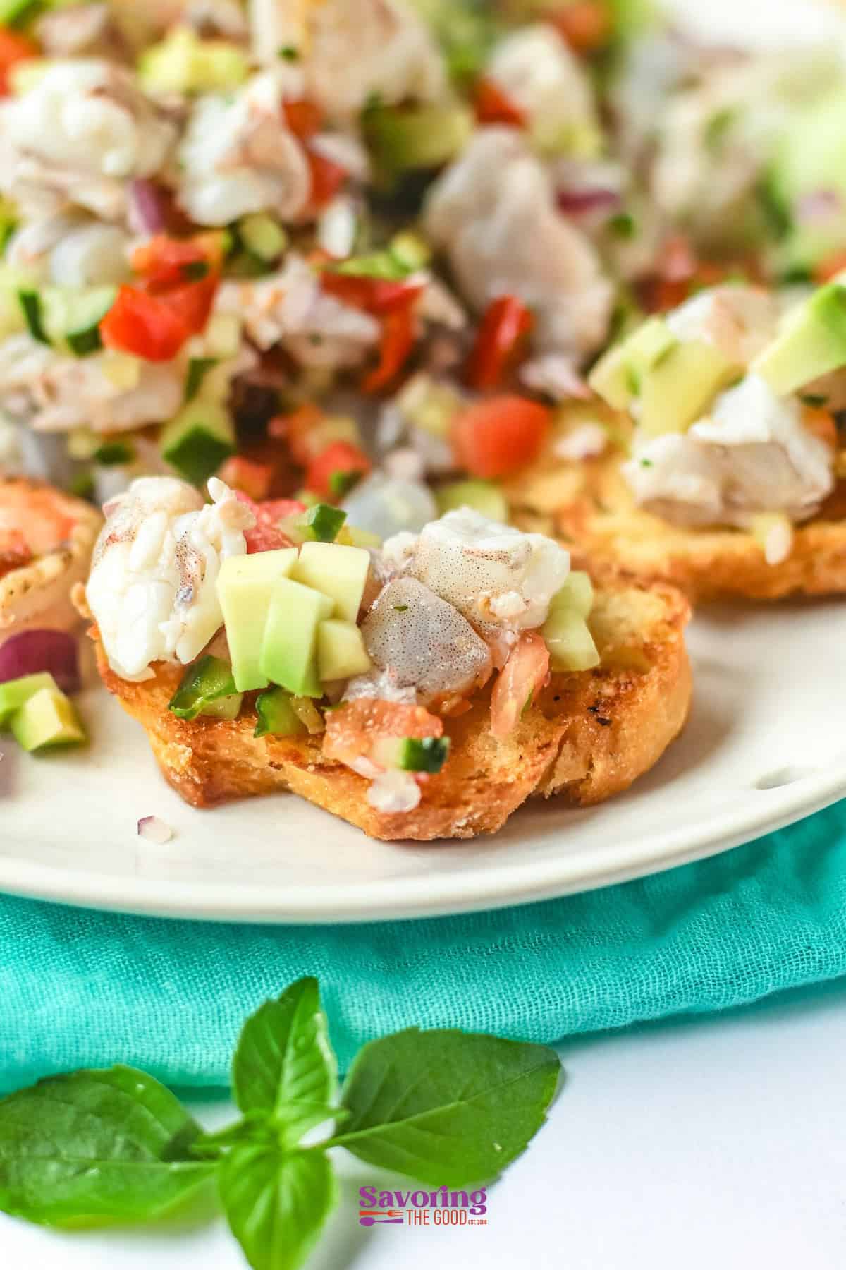 A close-up of seafood bruschetta topped with diced avocado, red onion, tomato, and herbs on a white plate with a teal napkin underneath.