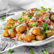 A plate of tater tots topped with chopped bacon, drizzled with sauce, and garnished with green onions. The plate sits on newspaper, with some toppings scattered around.