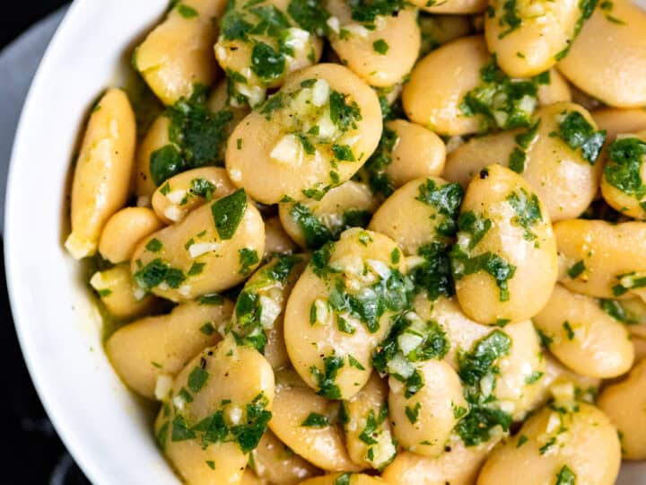 A white bowl filled with cooked butter beans mixed with chopped parsley, garlic, and olive oil-an ideal side if you're wondering what to serve with chicken drumsticks.
