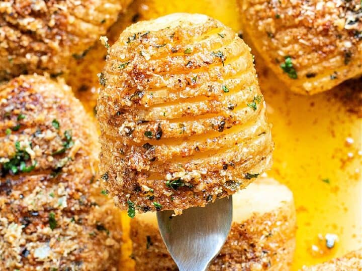 A close-up of hasselback potatoes topped with breadcrumbs and herbs, with one potato lifted by a fork above the rest in a baking dish with melted butter.