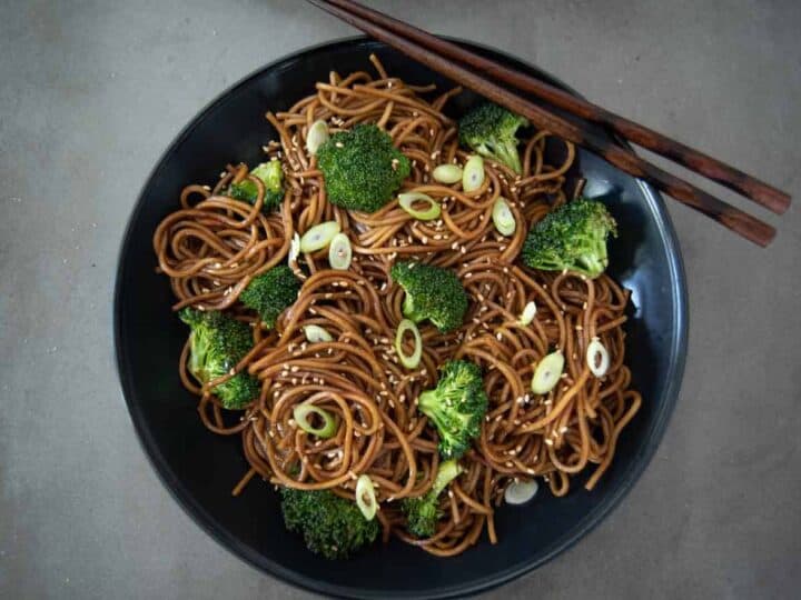 A black bowl of noodles with broccoli, sliced green onions, and sesame seeds sits ready to enjoy-an ideal option for what to serve with chicken drumsticks. A pair of chopsticks rests above the bowl on a gray surface.