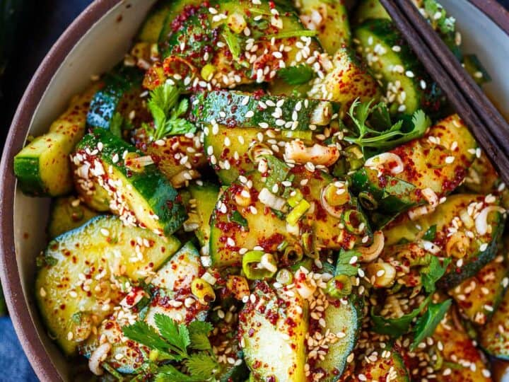 A bowl of sliced cucumbers topped with sesame seeds, chopped herbs, and red pepper flakes, with a pair of chopsticks resting on the bowl-an ideal side dish when wondering what to serve with chicken drumsticks.
