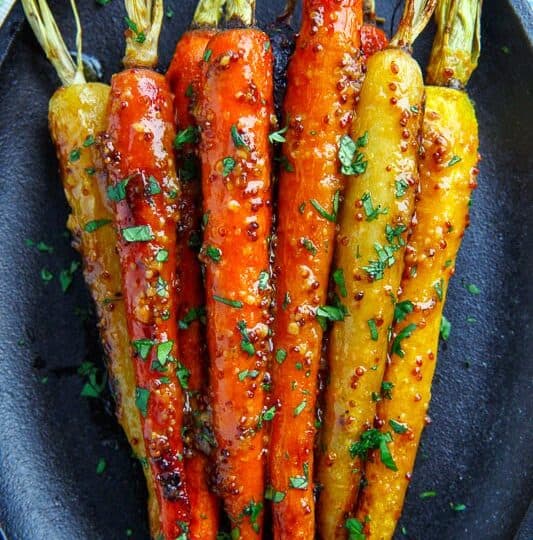 A black plate with roasted orange and yellow carrots garnished with chopped herbs and a visible mustard glaze-an ideal side when considering what to serve with chicken drumsticks.