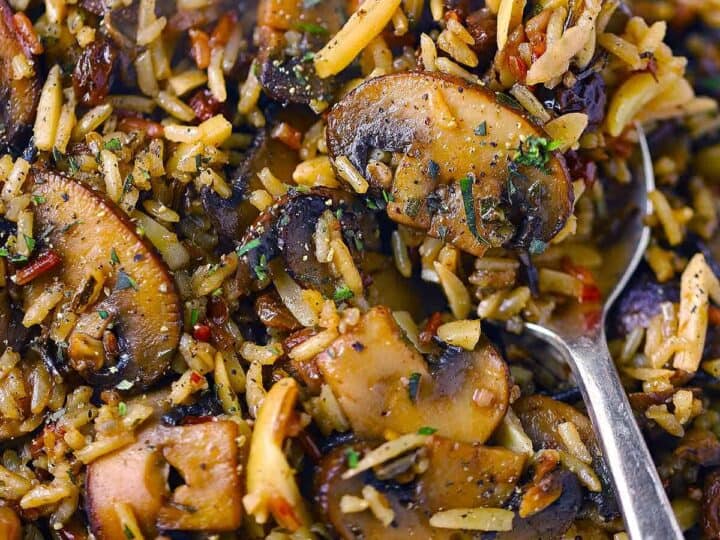 Close-up of a rice dish with sliced mushrooms, herbs, and visible spices, with a metal spoon partially in view-an ideal side when exploring what to serve with chicken drumsticks.