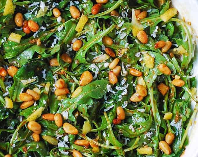 A bowl of arugula salad topped with toasted pine nuts, shaved Parmesan cheese, and a light vinaigrette, served on a wooden surface-an ideal side when wondering what to serve with chicken drumsticks.