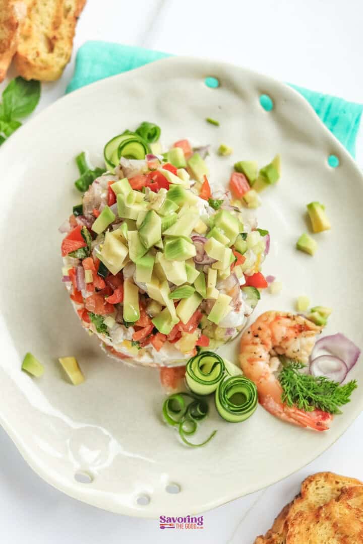 A white plate holds a shrimp ceviche topped with diced avocado, red onions, and tomatoes. A shrimp and cucumber garnish are on the side. Toasted bread is partially visible in the background.