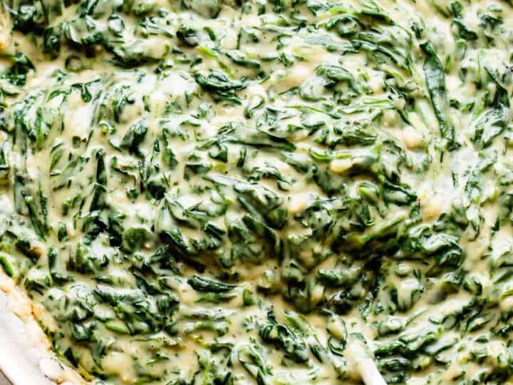 A close-up of creamy spinach in a skillet being stirred with a white spoon-a perfect side when deciding what to serve with chicken drumsticks.