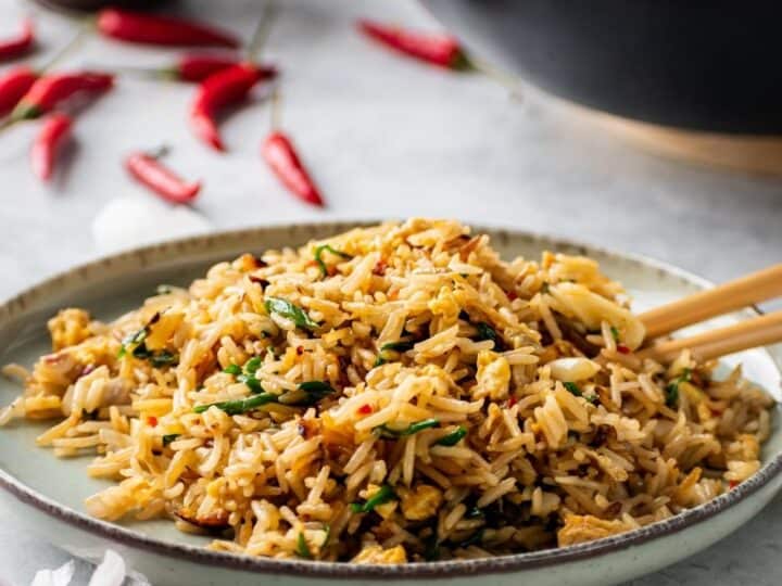 A plate of fried rice garnished with herbs, served with chopsticks. A wok, bowl of red chili peppers, and scattered garlic cloves are in the background-an ideal choice for what to serve with chicken drumsticks.