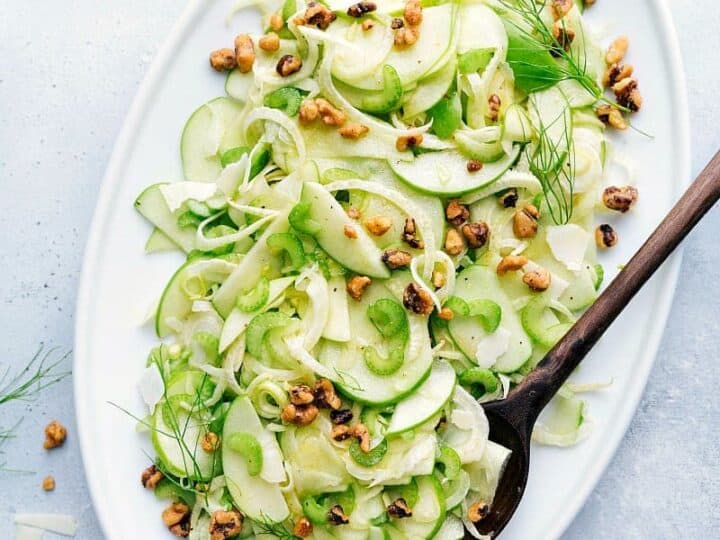 Oval white plate with thinly sliced cucumber and fennel salad, topped with walnuts, on a light marble surface. A wooden spoon rests on the edge of the plate-perfect for what to serve with sea bass.