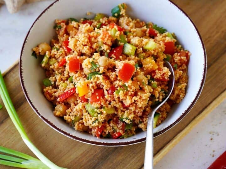 A bowl of vegetable couscous with diced bell peppers, zucchini, and chickpeas, garnished with green onions. Perfect for what to serve with sea bass, it's accompanied by a spoon, and a halved red pepper and scattered green onions resting on a cutting board.