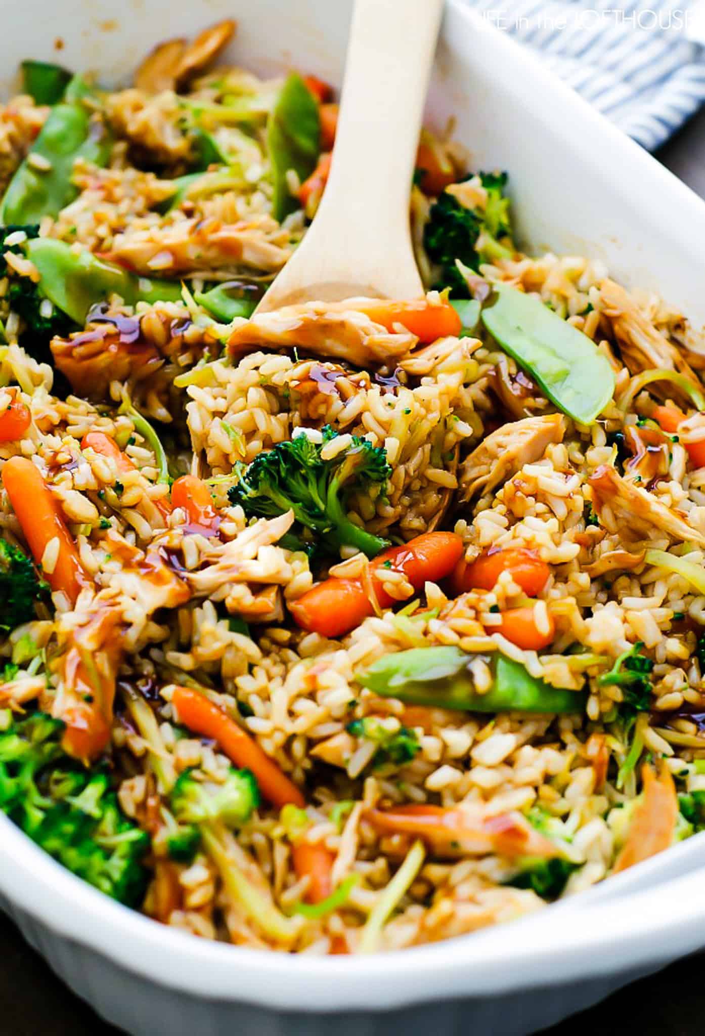 A close-up of a dish filled with mixed vegetables, rice, and chicken, topped with a dark sauce, with a wooden spoon in the dish. Perfect for anyone in search of hot dish recipes.
