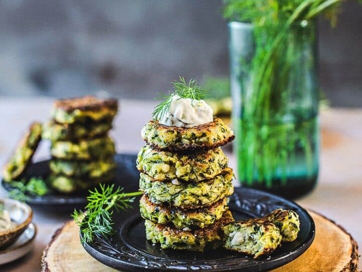 A stack of three zucchini fritters topped with a dollop of sour cream and a dill sprig, placed on a black plate with a wooden slab base, surrounded by more fritters, dill, and a glass vase with dill. These make an elegant side dish if you're wondering what to serve with sea bass.