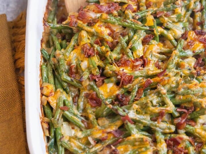 Casserole dish filled with green beans, melted cheese, and bacon, with a wooden serving spoon. Brown cloth napkin placed beside the dish. This savory casserole makes an ideal side when considering what to serve with sea bass.