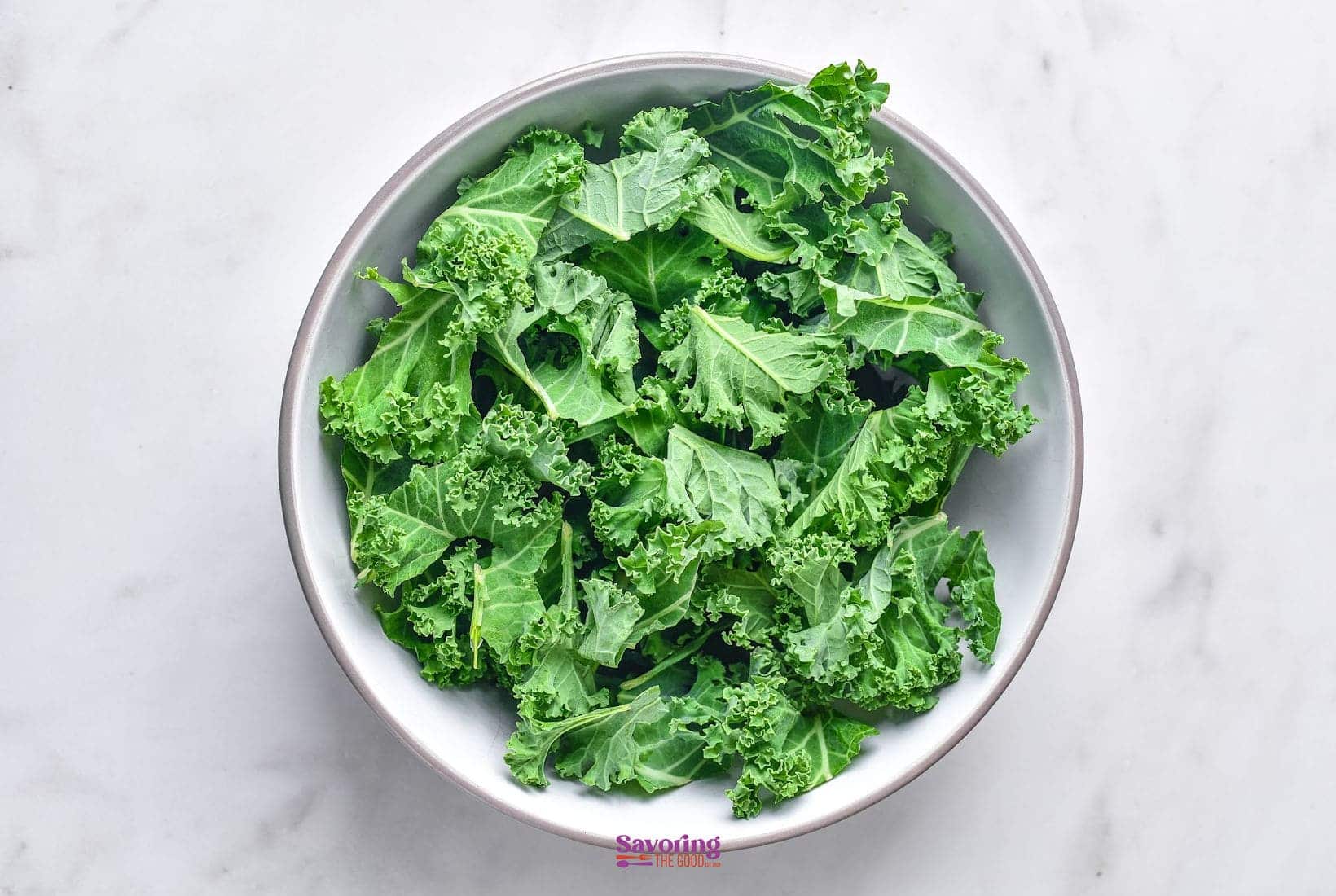 A white bowl filled with fresh, crisp kale sits on a pristine white marble surface.