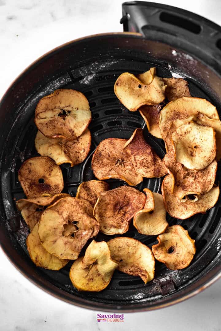 An air fryer basket filled with sliced, seasoned apple chips on a white background.