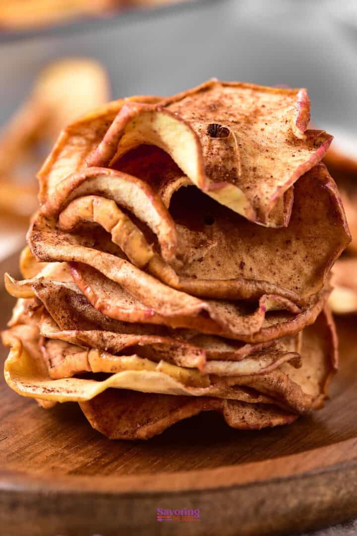 A stack of cinnamon apple chips on a wooden surface. The chips are thinly sliced, browned, and slightly curled, suggesting they have been baked or dehydrated.