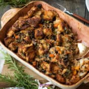 A casserole dish filled with a delicious vegetable stuffing recipe, containing chunks of bread, diced vegetables, and herbs, with a serving spoon placed on one side. Fresh rosemary sprigs are nearby.
