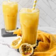 Two tall glasses of refreshing passion fruit juice with ice and striped straws sit on a marble surface. Nearby, halved passion fruit and a yellow cloth add vibrant color to the scene.