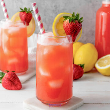 A glass of refreshing strawberry lemonade with ice, garnished with a lemon slice and whole strawberry, placed on a coaster. Other glasses, fresh strawberries, and lemons are in the background.
