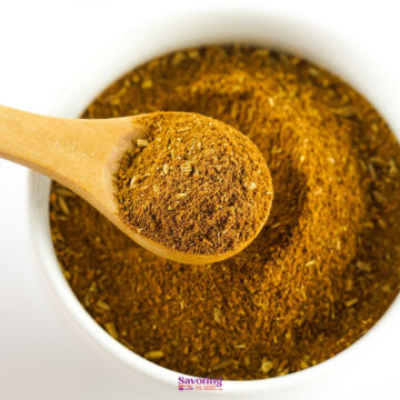 A wooden spoon filled with Chinese five spice is resting on the rim of a white bowl containing more of the same spice blend.