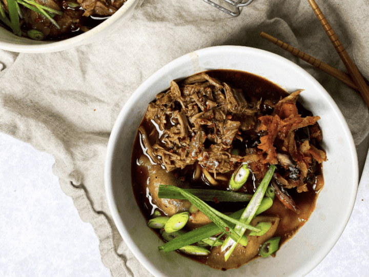 A bowl of beef ramen with green onions and other garnishes on a beige cloth, accompanied by chopsticks.