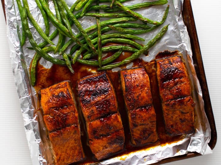 A baking sheet with four cooked salmon fillets, green beans, and lemon slices on foil. The salmon is seasoned and browned.
