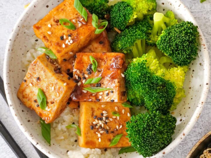 A bowl of rice topped with tofu cubes, broccoli florets, and garnished with sesame seeds and green onions. Black chopsticks are placed beside the bowl.