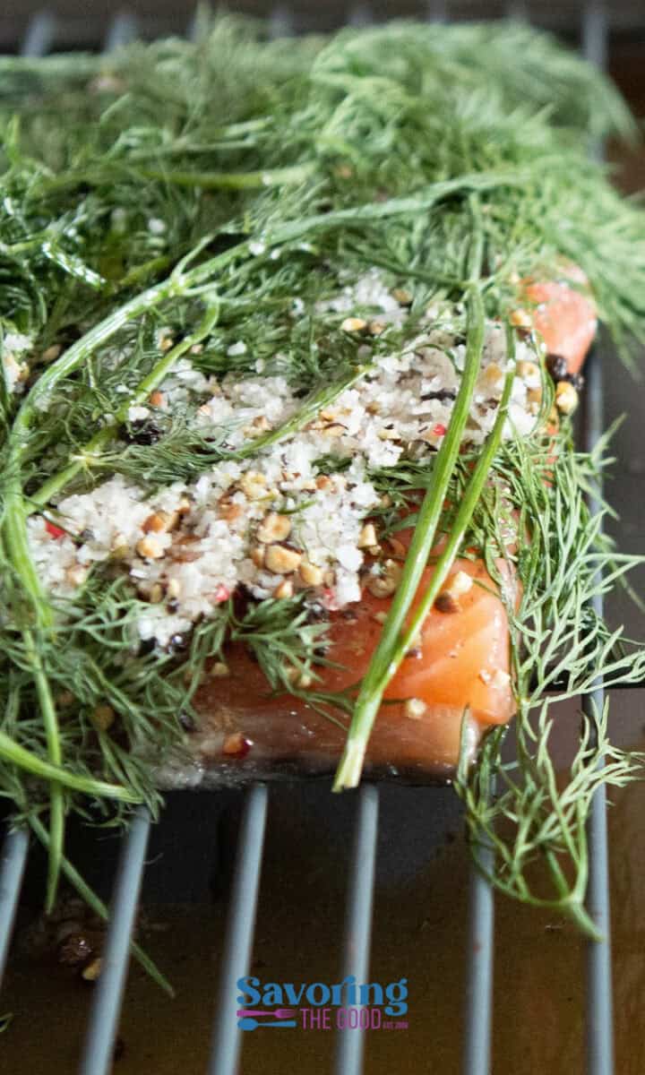 A piece of raw salmon fillet covered with dill, coarse salt, and spices is placed on a metal rack.