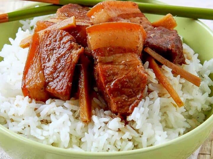 A bowl of white rice topped with pieces of braised pork and garnished with green chopsticks.