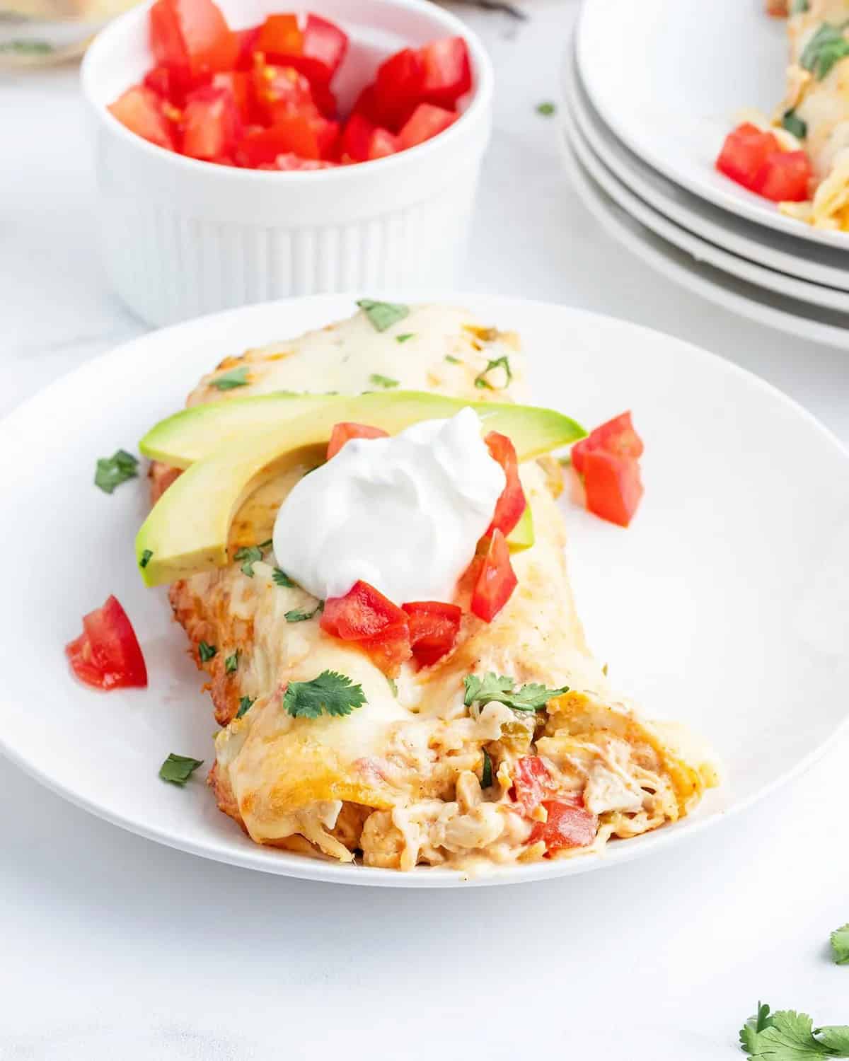 A plate features an enchilada topped with melted cheese, diced tomatoes, avocado slices, sour cream, and cilantro, with a bowl of diced tomatoes in the background.