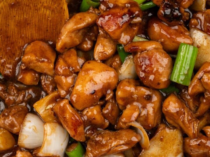 Close-up of a stir-fry dish featuring chunks of cooked chicken, green onions, and white onions, all coated in a rich brown sauce, with a wooden spatula on the side.