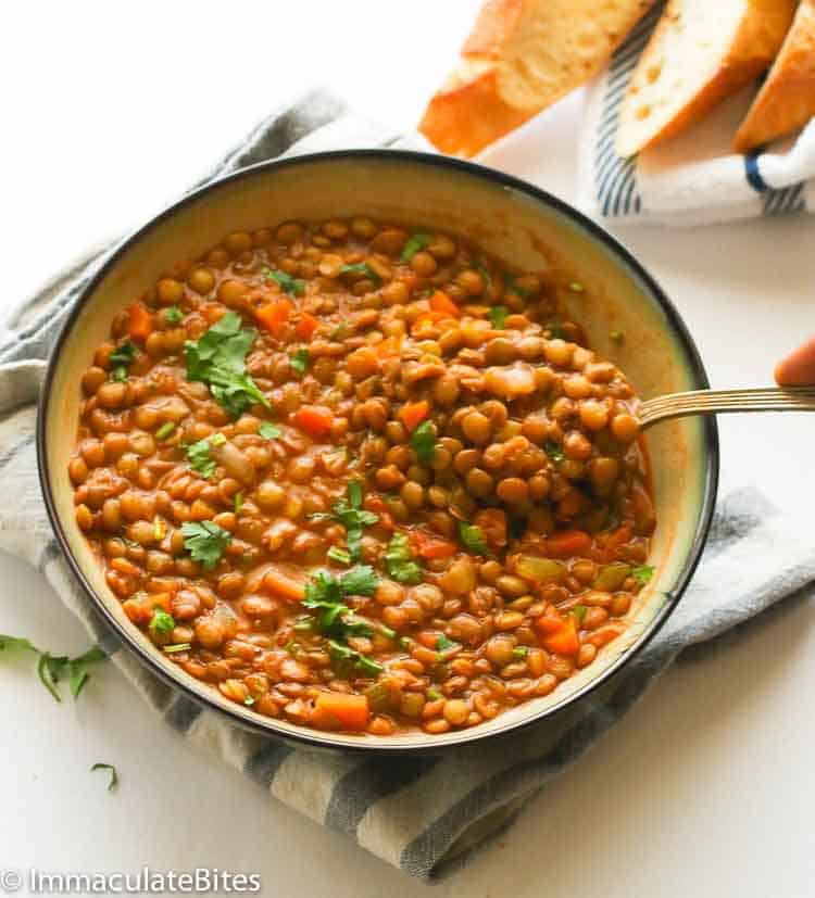A bowl filled with lentil stew garnished with fresh herbs, perfect for a cozy night in. A spoon is lifting some of the comforting stew, and slices of bread are visible in the background, making it one of those soup recipes you'll want to revisit.