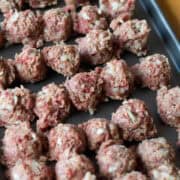 A baking sheet with rows of uncooked meatballs with chopped onions, placed on a wood surface.