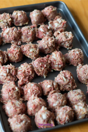A baking sheet with rows of uncooked meatballs with chopped onions, placed on a wood surface.